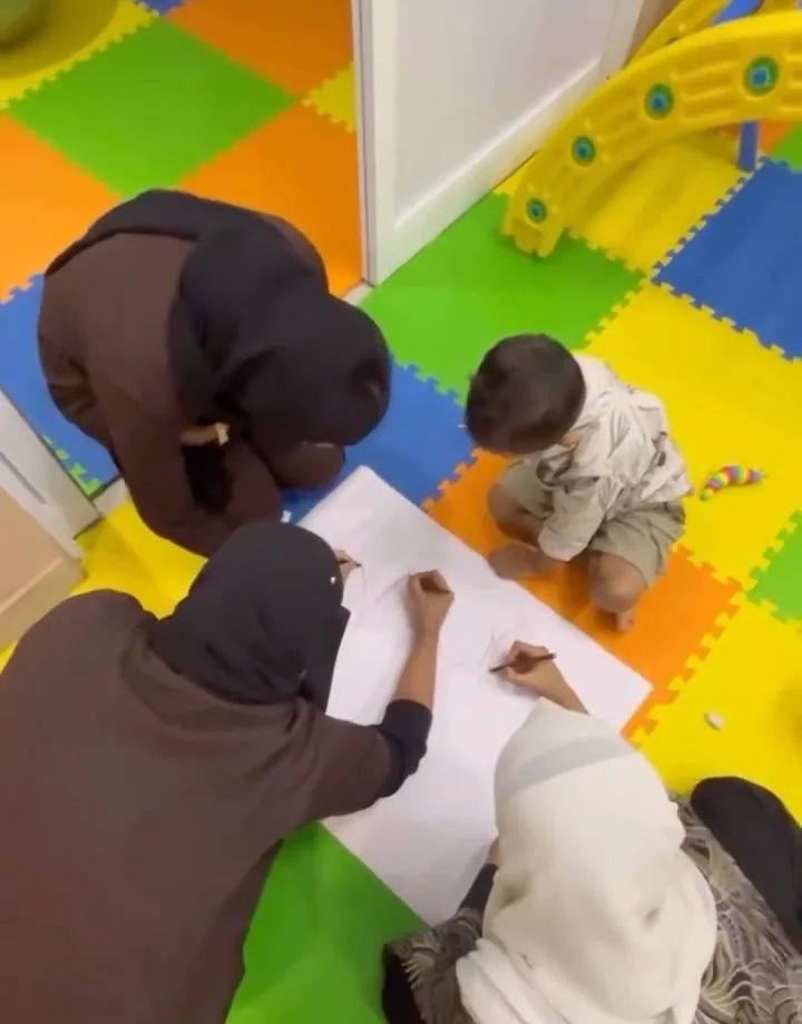 Therapists draw on a large sheet over bright foam play mats while a toddler sits beside them